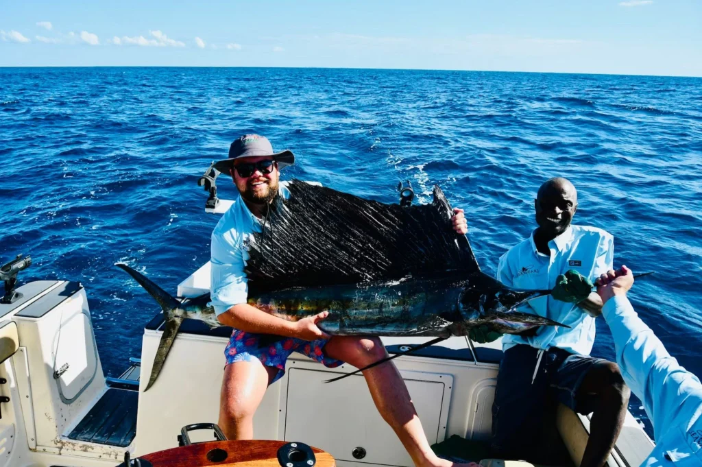 Two smiling men on a boat proudly holding a large sailfish they caught during a deep-sea fishing trip, with the open ocean and clear blue sky in the background.