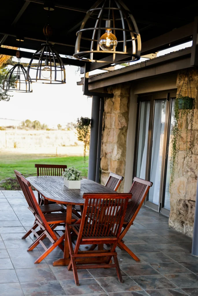 Outdoor covered patio with a wooden dining set and decorative hanging lights, overlooking a grassy field.