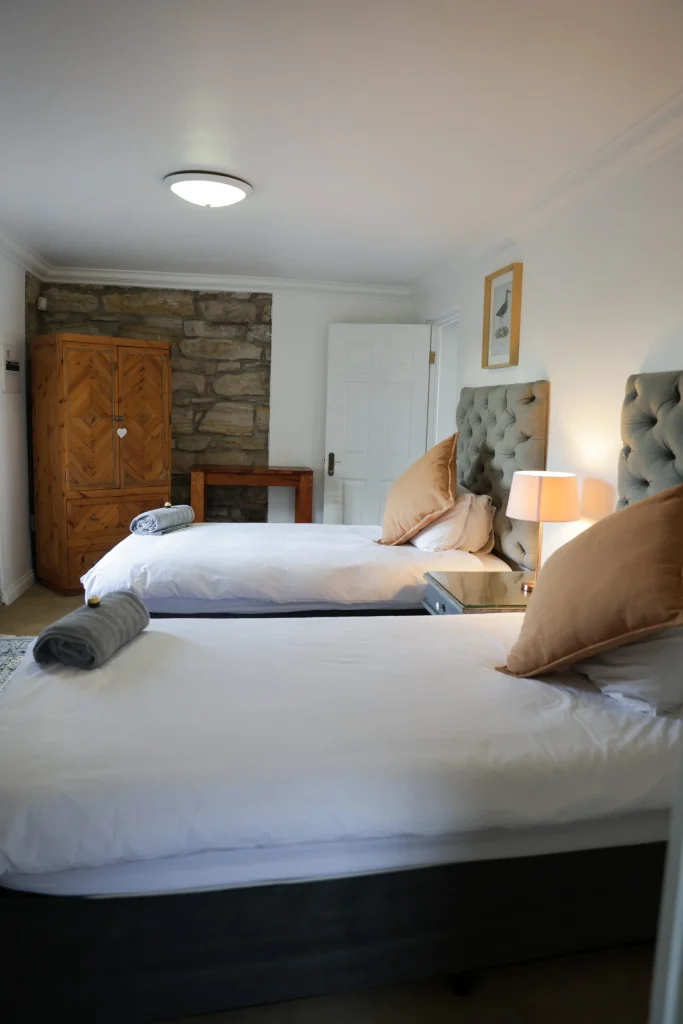 Bright twin bedroom with upholstered headboards, wooden wardrobe, and rolled towels placed neatly on each bed.