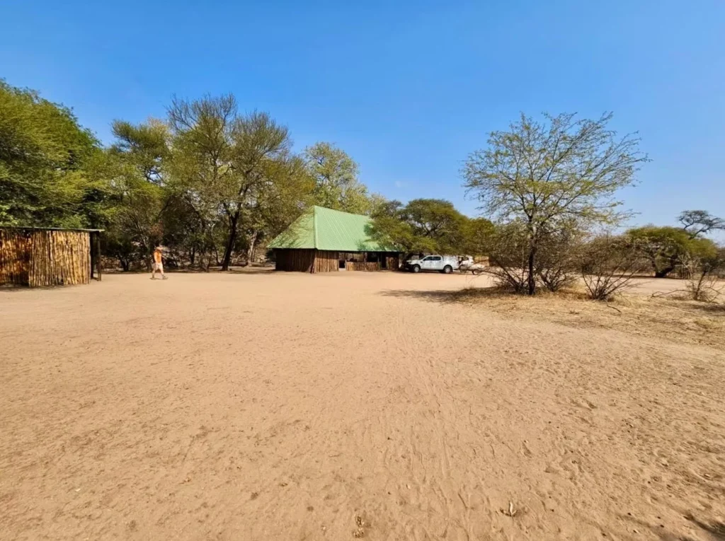 Wide view of a Botswana safari camp with sandy terrain, surrounding trees, and a central thatched building.